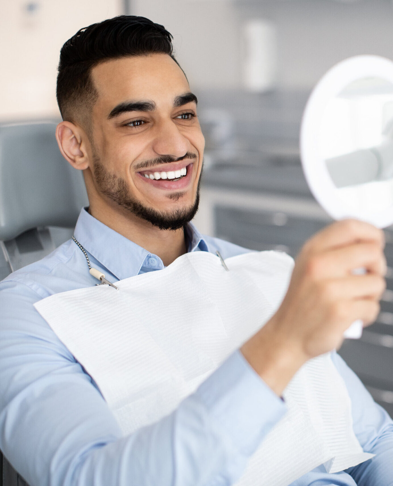 Happy Handsome Arab Man Looking To Mirror After Teeth Treatment In Modern Clinic, Middle Eastern Male Patient Sitting In Chair In Stomatological Cabinet And Enjoying His New Smile, Closeup